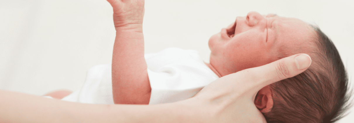 Crying newborn held by an adult's hand.